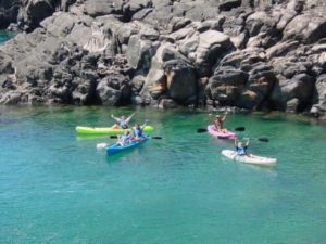 People kayaking in clear blue water near rocky cliffs.